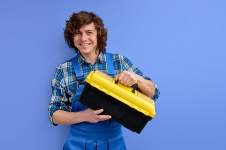 Young builder man in workwear holds tool box in hands and looks at camera smiling isolated over blue...