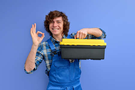 Man in coveralls hold tool case box isolated on blue studio background, showing OK gesture. Instruments accessories for renovation apartment room. Repair home concept. Curly caucasian smilingの写真素材