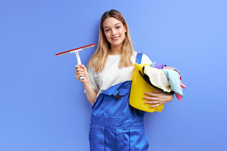 Smiling woman housewife in gloves holding cleanser things isolated over blue studio background....