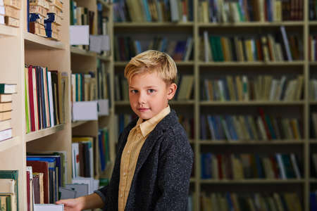 kid boy in library, looking at camera. school boy stand between shelves with booksの写真素材