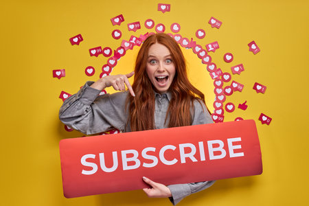 Redhead female asking recommending to subscribe, pay attention, holding title in hands, pointing index finger. Social media concept. Isolated yellow backgroundの写真素材