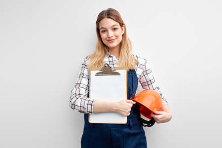 Confident cheerful woman engineer holding paper tablet and helmet, dressed in builder uniformの写真素材