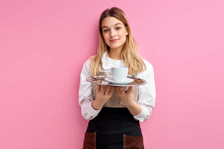 Beautiful waitress offering cup of coffee isolated on pink backgroundの写真素材