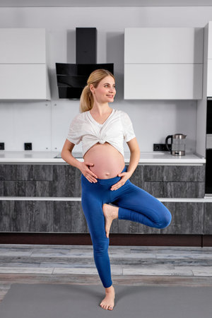 Full-length portrait of young female in sportswear standing on one leg stroking tummy, meditating