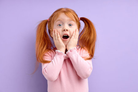 nice little surprised girl with ginger red hair and freckles, stands over purple background touching...