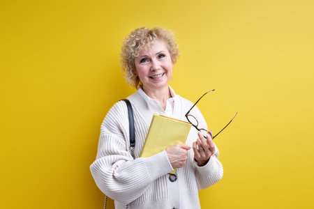 Open-minded senior woman holding book and eyeglasses in hands, confident teacher ready to teach you,...