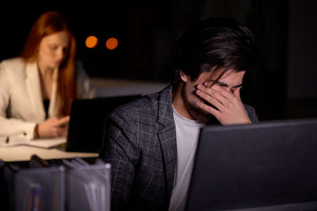 Tired and overworked business man manager sitting in front of computer laptop and trying to concentrate. male worker eyes are dry and hurt painful. young guy work in night officeの写真素材