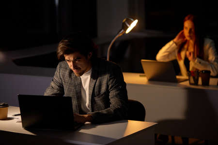Portrait of tired focused young man working remotely late at night in dark office using computer, typing, thinking about deadline, in formal wear. Overwork, negative thoughts, deadline conceptの写真素材