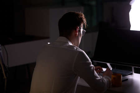 Unrecognizable Male Programmer Writing Code on Computer at Night. Side View. Young Guy In Formal Wear Engaged In Work, In Office In Dark Office. Networking, Freelance, Deadline Conceptの写真素材