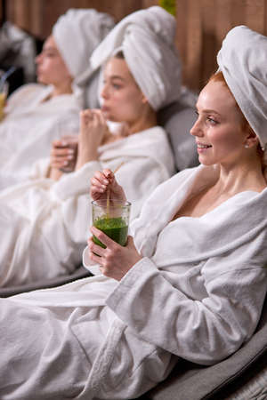 Caucasian women relaxing and drinking juicy beverage smoothies in robes during wellness weekend in wellness center, lying on bed, after spa procedures. focus on redhead female with towel on headの写真素材