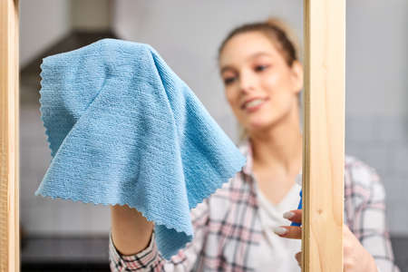 Female cleaning window glass with blue rag. Close-up photo of rag. Spring cleanup, housework concept. Focus on blue cloth, smiling woman in the backgroundの写真素材
