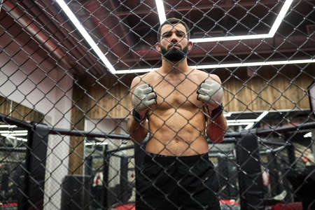 Portrait of MMA fighter thinking about strategy for match, standing on ring preparing for fight, shirtless muscular male is going to practice fighting. shot through cageの写真素材