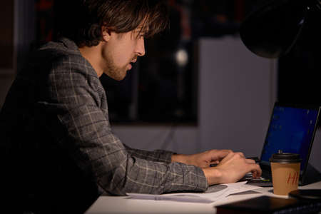 Confident Male Programmer Writing Code on Computer at Night. Table Light Turned On. Caucasian Guy In Formal Wear Engaged In Work, In Office In Dark Office. Networking, freelance, deadline. Side Viewの写真素材