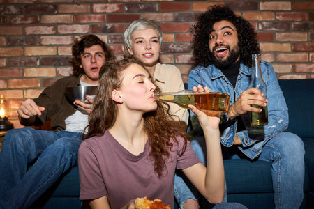 young friends watching tv, comedy show or movie, eating snacks and drinking beverage, sitting on cozy couch at home, diverse guys and ladies enjoying free time, weekend togetherの写真素材