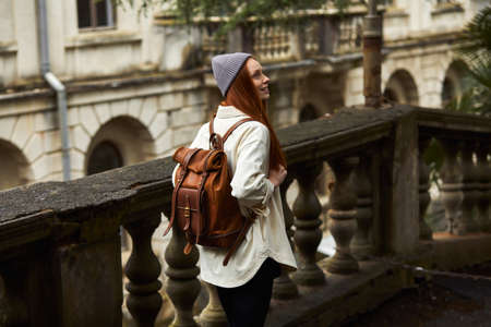 Rear view on redhead woman in coat in search of beautiful historical place, looking around, stand in contemplation, exploring new area, young traveler enjoy walking alone. travel, trip conceptの写真素材
