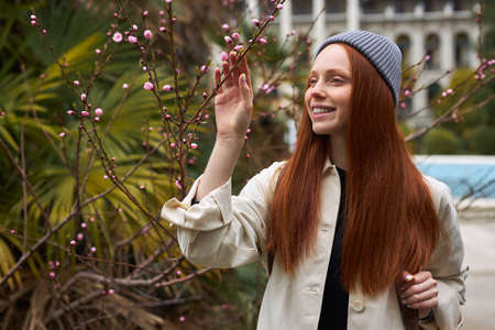 Young woman in spring garden. Springtime. Trendy redhead female enjoy feeling smell of blooming flowers on tree, looking at side, smiling, walking alone in good mood, building in the backgroundの写真素材