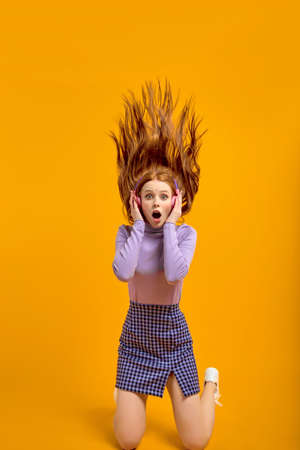 Caucasian Funny Female With Flying Long Red Hair Is In Shock By Listening Music So Loud, Look At Camera With Surprised Facial Expression, Opening Mouth By Shock, Isolated On Bright Yellow Backgroundの写真素材