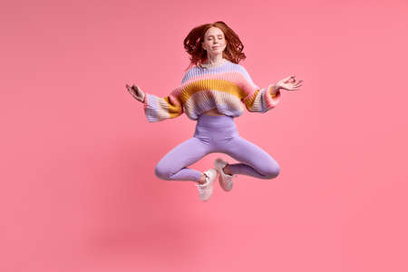 beautiful redhead lady jumping high sitting air in lotus pose practicing morning yoga wearing casual outfit isolated pastel pink color background, portrait. Calm caucasian female meditatingの写真素材
