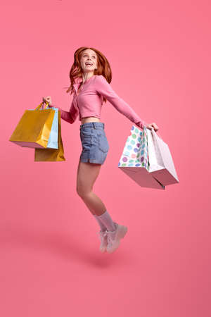 Crazy beautiful redhead lady jump high up speed rushing black friday addicted shopper wearing pink shirt and denim shorts, carrying many packages, isolated pink pastel color backgroundの写真素材