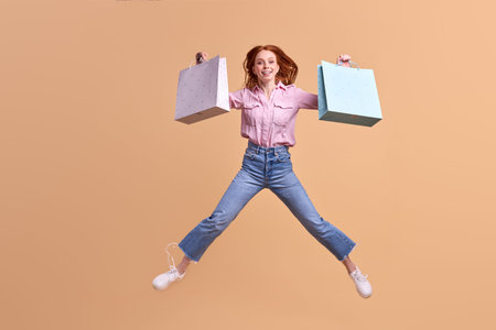 Happy red-haired lady happy after shopping, carry packages, in casual modern stylish denim jeans, jumping, having fun. Isolated pastel yellow background. copy spaceの写真素材