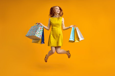 Full length photo of pretty redhead lady carry many shopping packs, addicted shopaholic in yellow dress, rejoicing shopping center isolated ove yellow color background in studioの写真素材