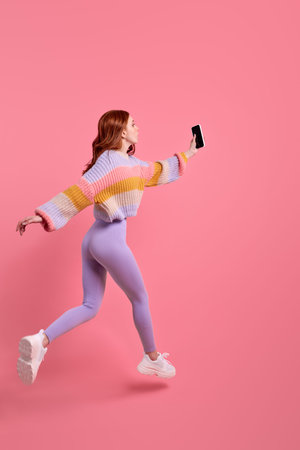 side view portrait of redhead female in leggins running jumping forward with smartphone in hands,...