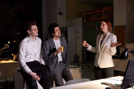 Side view on friendly office workers, colleagues having talk after hard working day, at night. Redhead woman and two guys in formal wear discussing, taking a break, bussiness team in boardroomの写真素材