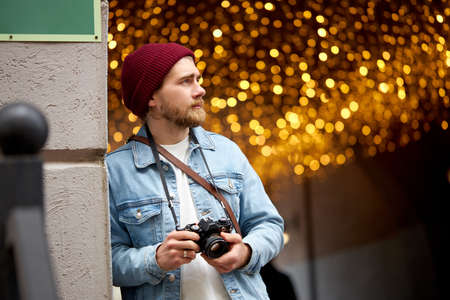 Caucasian hipster guy in denim jacket and red hat walk in city street taking photo, using film camera, Garlands lights in the background, copy space. Photography, people lifestyle concept.の写真素材