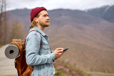 caucasian brutal bearded man traveling in the wild nature using smartphone, with leather brown...