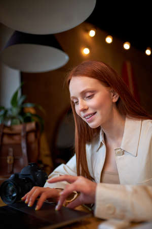 Positive redhead female designer working on marketing project at cafe, skilled female photographer noting ideas earning money on editing images on laptop, side view portraitの写真素材