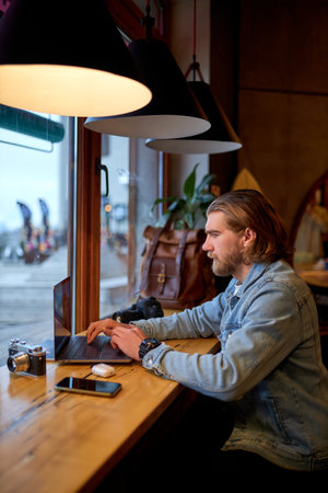 Portrait of concentrated bearded male photographer journalist sitting at cafe working with digital...