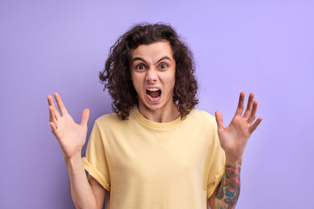 Nervous irritated curly man scream at camera spreading arms, feeling stress, worried about something, stand with wide opened mouth isolated over purple backgroundの写真素材
