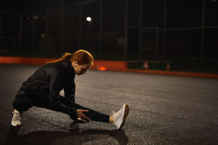 Motivated redhead female athlete stretching legs for warming up before urban fitness and running workout at night in city, on road. Sporty woman exercising outdoors, side view portrait. copy spaceの写真素材