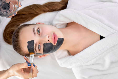 Hands of cropped Caucasian cosmetologist woman applying facial mask on beautiful young Caucasian womans face lying on massaging bed,top view portrait. copy space. beauty, skin care, spa conceptの写真素材
