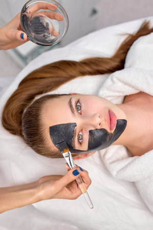 Procedure for applying black mask on fresh skin of face of beautiful woman, top view. Spa...