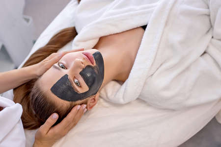 Young caucasian female uses the services of professional beautician at at spa. cropped unrecognizable master beautician puts black mask on clients face and gives a massage. Health and longevityの写真素材
