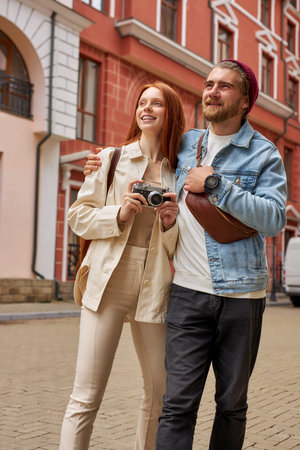Caucasian tourists man and woman taking photo of architecture on retro film camera, wearing casual...