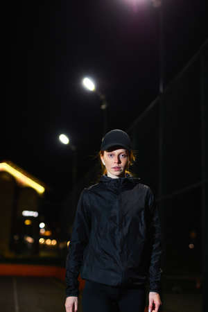 pretty young serious redhead female in black sportive clothes posing looking at camera outdoors, after jogging running, sport fitness training. at night in city alone. copy space. people lifestyleの写真素材