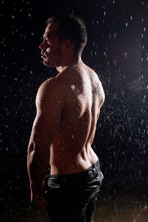 Side view on sexy muscular sportive man with strong body, showing muscular back standing isolated against black background, in rain, water drops on body. Caucasian european male modelの写真素材
