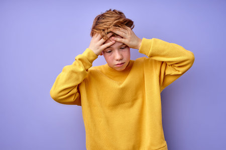 Teenage boy is suffering from headache, touching head, isolated on purple background. Portrait of...