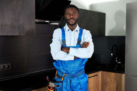 Serious Handsome Young African Handyman Plumber In Blue Uniform. Young Skilled Handyman In Overall Standing At Home In Kitchen Posing Looking At Camera. Repairman Service, Hard Work Conceptの写真素材