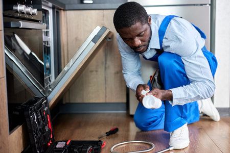 Side View Young Black Afro Handyman Repairman Repairing Dishwasher, Changing Siphon, Wearing Blue Workwear Overalls. Confident Professional Handyman ,Focused On Work Alone, In Kitchenの写真素材