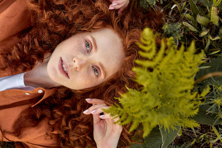 Young redhead curly female of caucasian appearance lying on grass in summer, posing looking at camera. attractive lady with natural beauty is looking dreamy and romantic . view from aboveの写真素材
