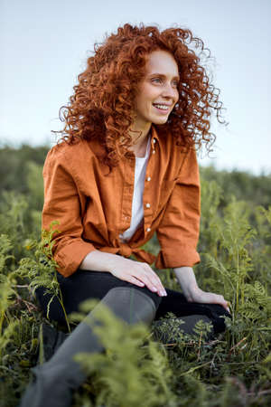 Smiling Lady Sitting On Green Grass In Field In Contemplation Of Beautiful Nature Around, Landscape. Redhead Curly Woman In Boots, Casual Shirt And Trousers Have Rest, Relaxing, Looking At Sideの写真素材