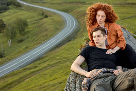 Exhausted Caucasian Young Couple Sitting On The Head Of Mountain, Having Rest After Climbing, Looking At Side In Contemplation of Nature Landscape Around. Road In The Backgroundの写真素材