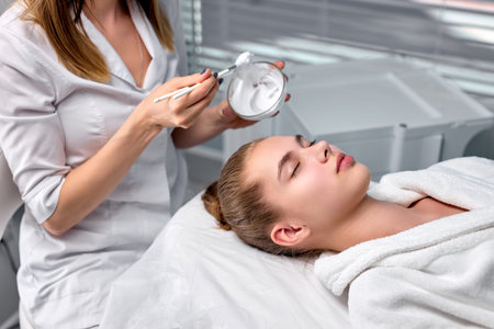 cropped cosmetologist applying mask from bowl on pretty woman face, gorgeous caucasian woman in spa...