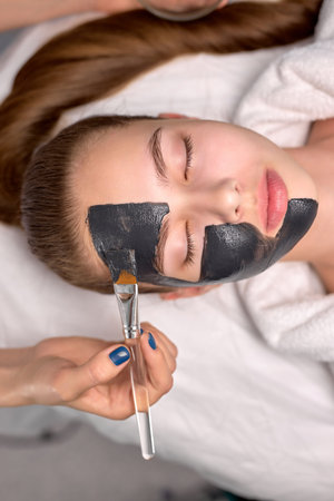 Top View On Cropped Professional Cosmetologist applying black mask on fresh face of beautiful woman...