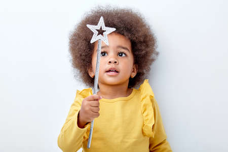 Cute african american girl wth curly fluffy hair holding magic wand, isolated on white studio wall. Funny little black girl wearing yellow casual shirt. Childhood lifestyle conceptの写真素材