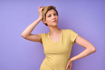 Confused short haired young female touching head having dilemma difficult choice dressed in casual yellow t-shirt isolated over purple studio background. Questioned female model. copy spaceの写真素材