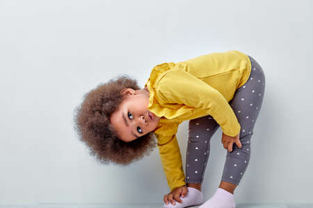 Charming Kid Girl Of African Appearance Stand With Head Bowed Looking Up, Having Perfect Charming Look And Fluffy Wavy hair, Dressed Casually. Pretty Kid Leaned On One Leg. Children Conceptの写真素材